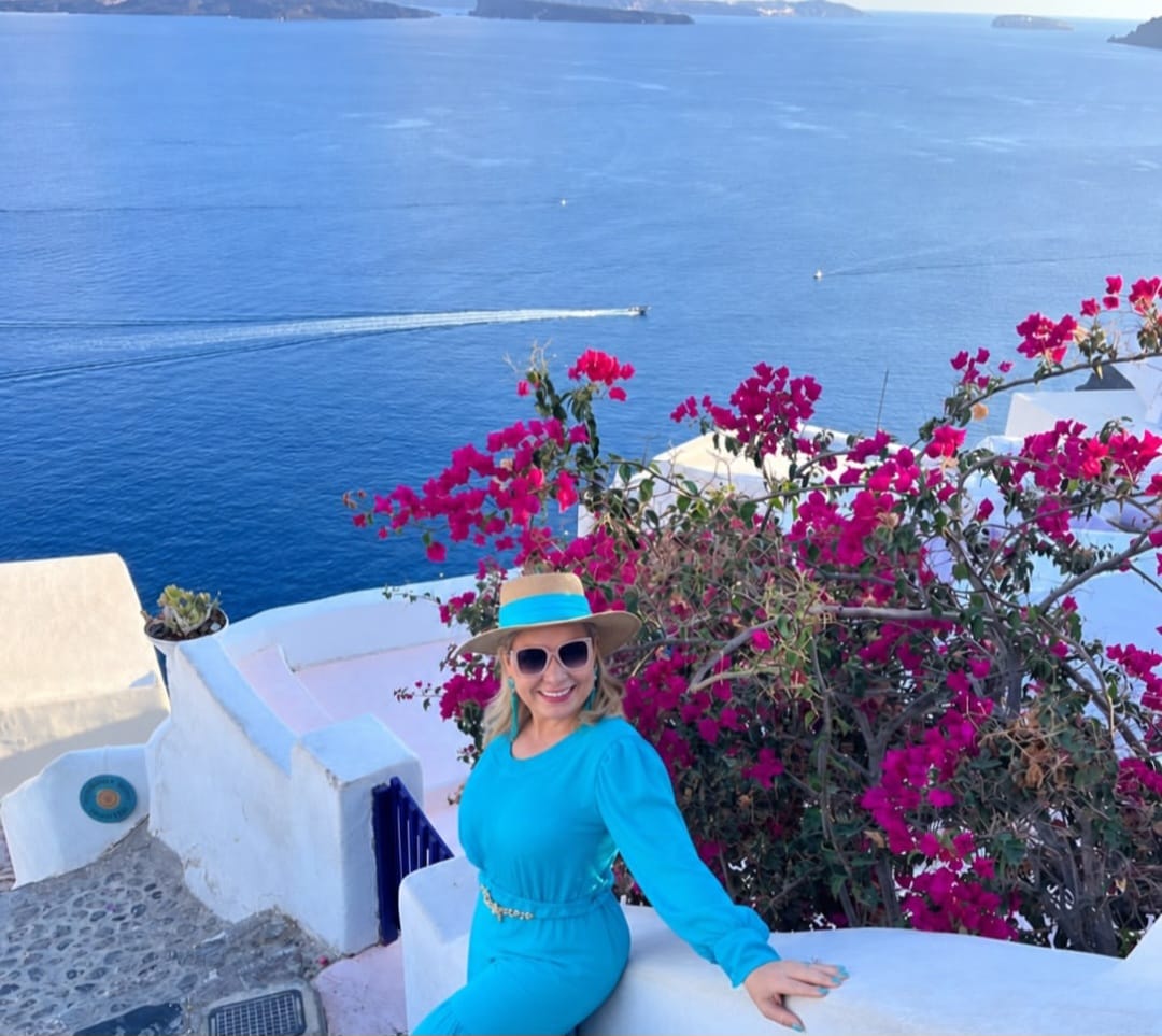 Sandra Martins de vestido verde-água e chapéu ao lado de buganvílias, com o mar ao fundo
